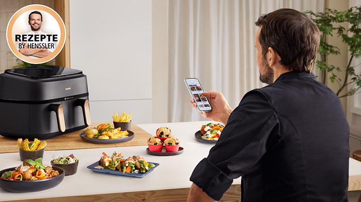 Steffen Henssler in schwarzer Kochjacke hält ein Smartphone auf dem eine App mit Rezepten geöffnet ist. Auf dem Tisch im Hintergrund sieht man unterschiedliche Speisen und einen Airfryer. In der Ecke ist ein Icon mit der Schrift "Rezepte by Henssler". 