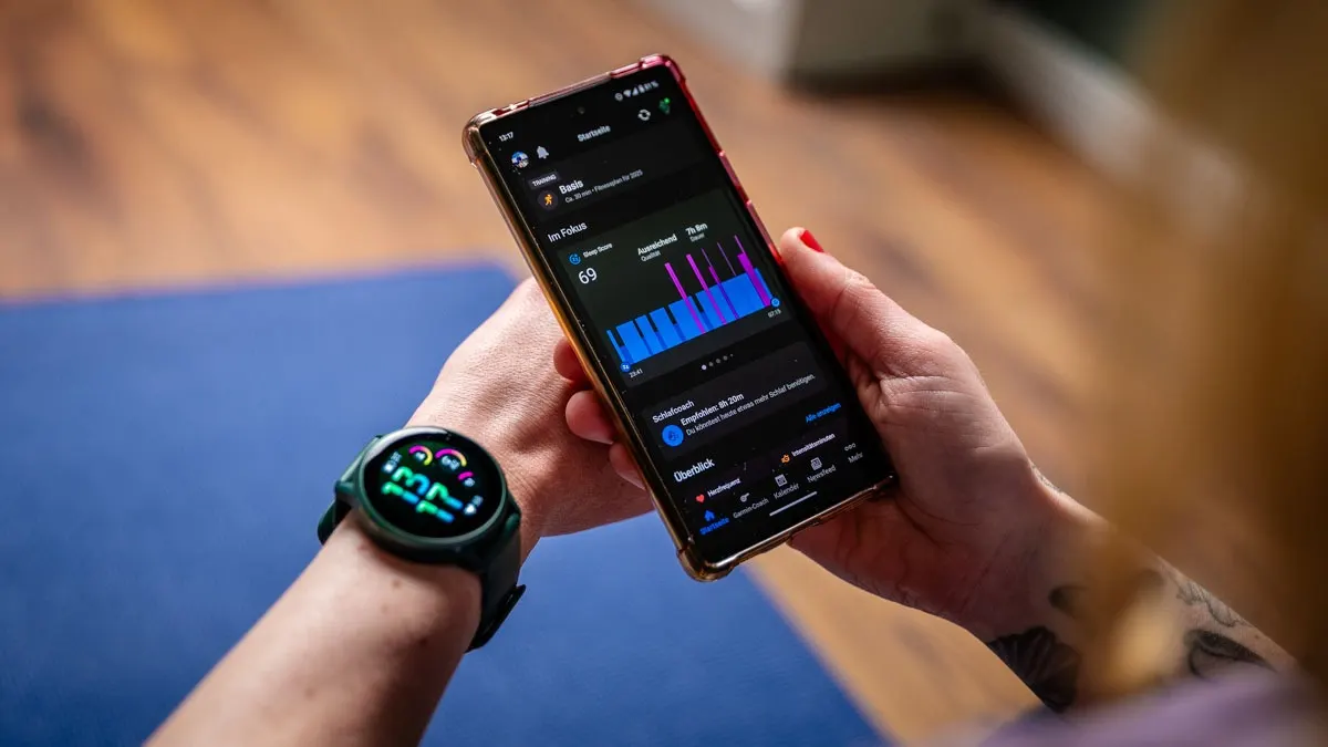Eine Person hält ein Smartphone mit einer Fitness-App und trägt eine Smartwatch am Handgelenk. Blaue Yogamatte im Hintergrund.