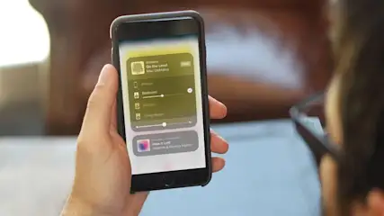 Ein Smartphone mit einer gelben App zur Lichtsteuerung wird in der Hand gehalten. Im Hintergrund sind Möbel zu sehen.