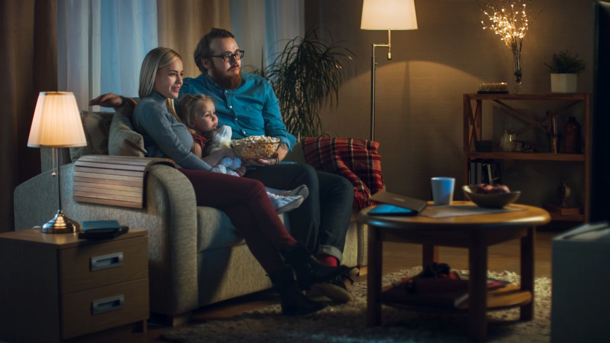 Eine Familie sitzt auf einem Sofa und schaut fern, der Mann hält eine Schüssel Popcorn. Ein Tisch und Regale sind im Hintergrund.