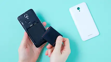 Hand hält Batterie neben offenem Smartphone; weiße Rückseite liegt daneben auf türkisfarbenem Grund.