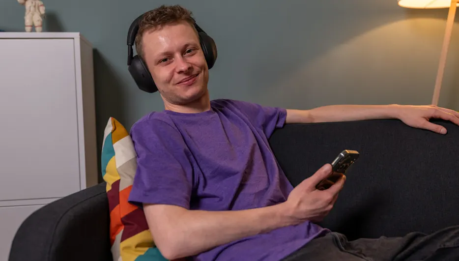 Alex sitzt auf einem Sofa und trägt den Kopfhörer Sony WH-1000XM6. In der Hand hält er ein Handy.