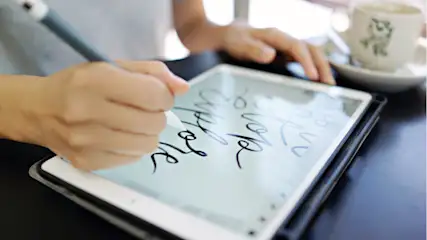 Eine Person schreibt mit einem Stift auf einem Tablet, daneben steht eine Tasse. Der Tisch ist dunkel.