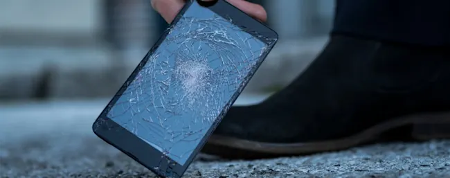Ein schwarzes Smartphone mit zerbrochenem Display wird von einer Hand gehalten, daneben steht ein schwarzer Schuh auf grauem Untergrund.