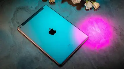 Auf grauem Grund liegt ein blaues iPad mit schwarzem Apple-Logo. Weiße Blüten sind oben rechts sichtbar, pinkfarbenes Licht rechts.
