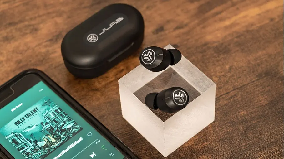 Schwarze Ohrhörer in durchsichtigem Würfel, daneben ein Etui und ein Smartphone mit Musik-App auf einem Holztisch.