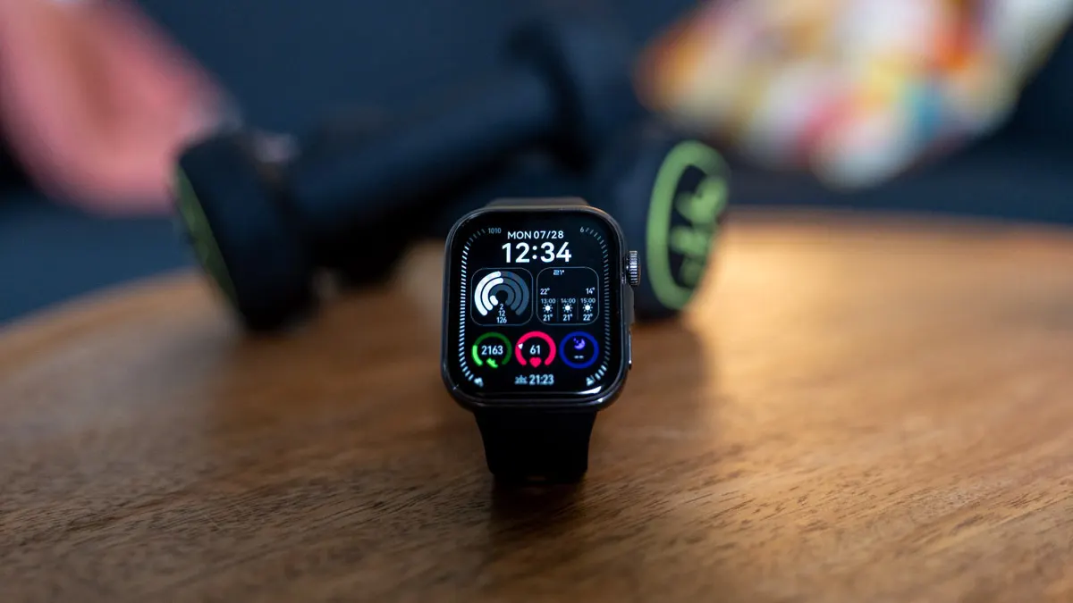 Die Huawei Watch Fit 4 steht aufrecht auf einem Tisch mit Hanteln im Hintergrund.