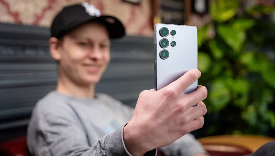 Mann mit Kappe hält silbernes Smartphone mit mehreren Kameras in der Hand, im Hintergrund Pflanzen und gemusterte Tapete.