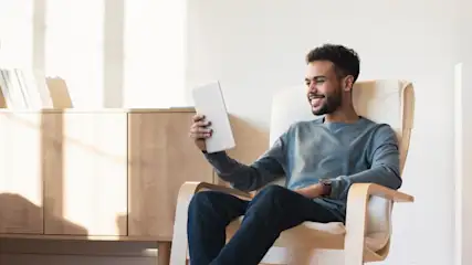 Mann sitzt in einem Sessel, hält ein Tablet in der Hand und lächelt. Im Hintergrund stehen ein Schrank und Bücher.