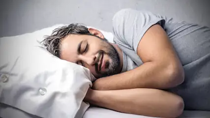 Ein Mann schläft auf einem weißen Kissen mit Knöpfen. Er trägt ein graues T-Shirt und liegt auf einem weißen Bettlaken vor einer grauen Wand.