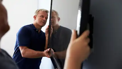 Ein Mann im blauen Poloshirt befestigt mit einer zweiten Person einen Fernseher an der Wand.