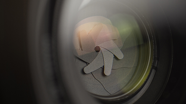 Nahaufnahme einer Kameraobjektivblende mit grauen Lamellen und einem gelb getönten Hintergrund.