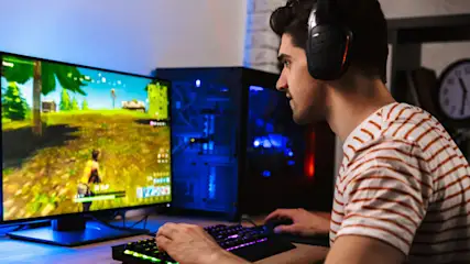Ein Mann mit Kopfhörern spielt ein Videospiel auf einem Computer mit beleuchteter Tastatur und Monitor. Im Hintergrund befindet sich ein PC-Turm.