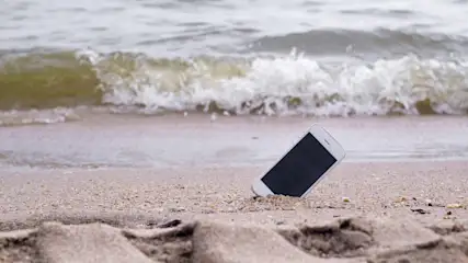 Ein weißes Smartphone mit schwarzem Bildschirm liegt am Strand, mit Wellen im Hintergrund.