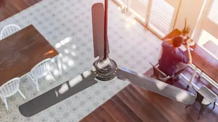 Deckenventilator mit drei Flügeln über einem Raum mit Tisch, Stühlen und einer Person am Fenster.