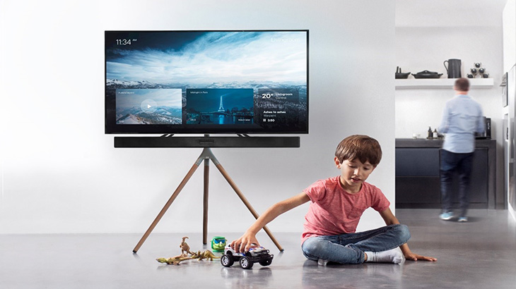 Ein Junge sitzt vor einem Fernseher auf einem Stativ und spielt mit Spielzeugautos. Im Hintergrund steht eine Person in der Küche.