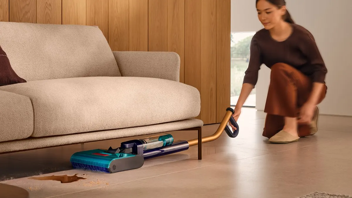 Eine Frau saugt mit dem Dyson Clean+Wash Hygiene Schmutz und eine braune Flüssigkeit unter einem Sofa auf.