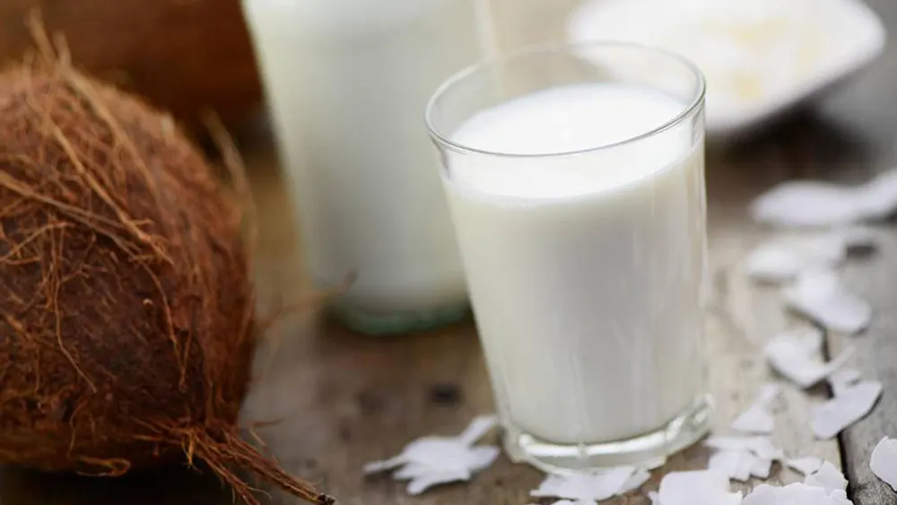 Eine Kokosnuss und ein Glas weiße Flüssigkeit stehen auf einem Tisch, mit Kokosraspeln drumherum.