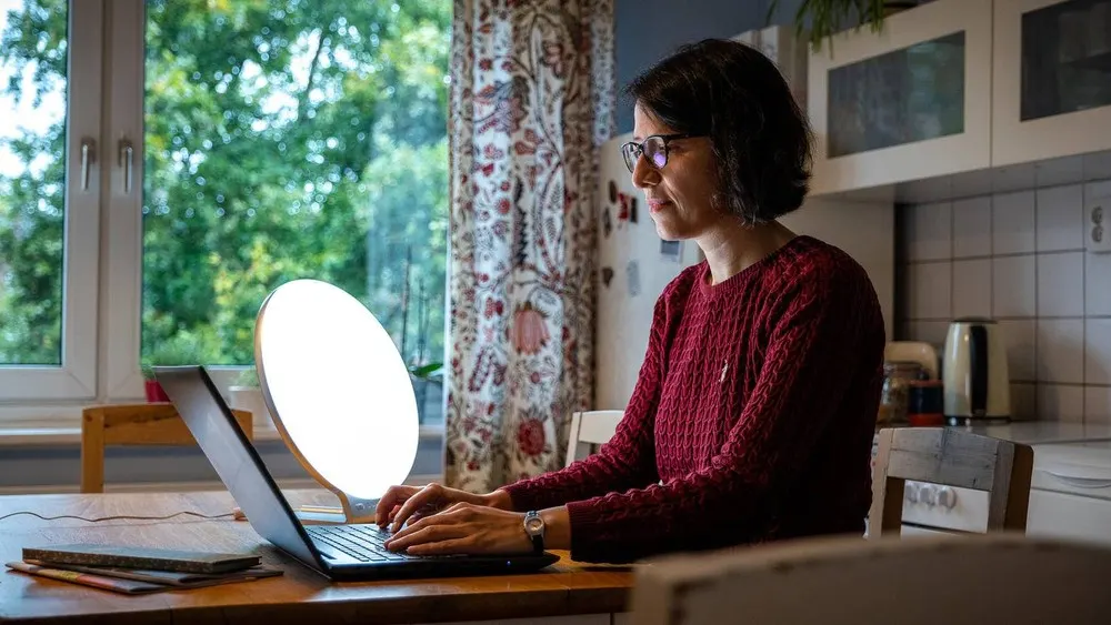 Frau mit Brille tippt auf Laptop an Tisch mit Lampe. Fenster, Vorhang und Grün im Hintergrund. Küche mit Schränken sichtbar.