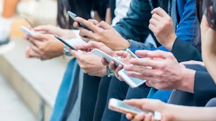 Mehrere Personen sitzen nebeneinander und halten Smartphones in ihren Händen. Einige tragen formelle Kleidung wie blaue Blazer.