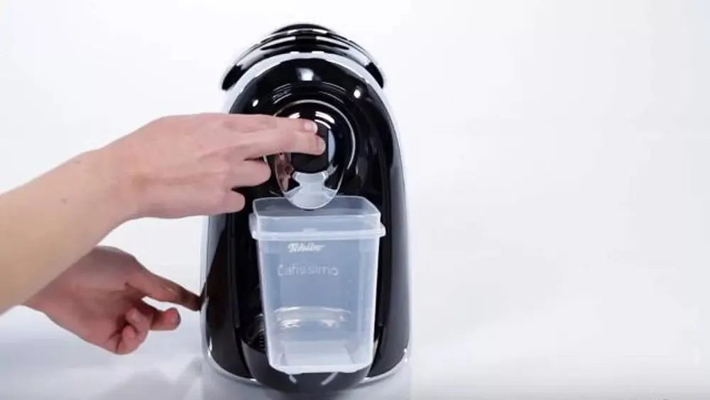 Eine Hand drückt einen Knopf an einer schwarzen Kaffeemaschine. Ein transparenter Behälter ist darunter befestigt. Weißer Hintergrund.