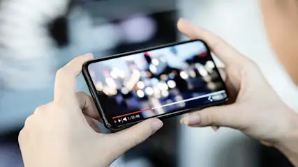 Eine Person hält ein Smartphone horizontal in beiden Händen, auf dem ein Video mit Wiedergabesteuerung läuft.