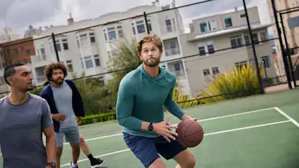 Drei Männer auf einem Basketballplatz. Einer hält einen Basketball, im Hintergrund Gebäude und ein Netz.