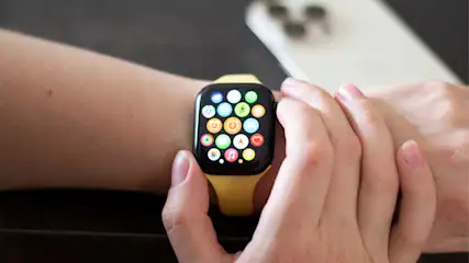 Eine Person bedient das App-Menü einer Apple Watch mit gelbem Armband. Im Hintergrund liegt ein weißes iPhone auf dem Tisch.