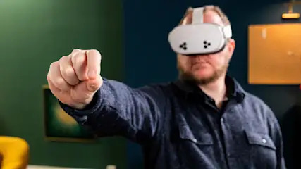 Mann mit VR-Brille und blauem Hemd reckt Faust vor grün-blauem Hintergrund. Ein gelber Sessel und ein Bild sind im Hintergrund sichtbar.