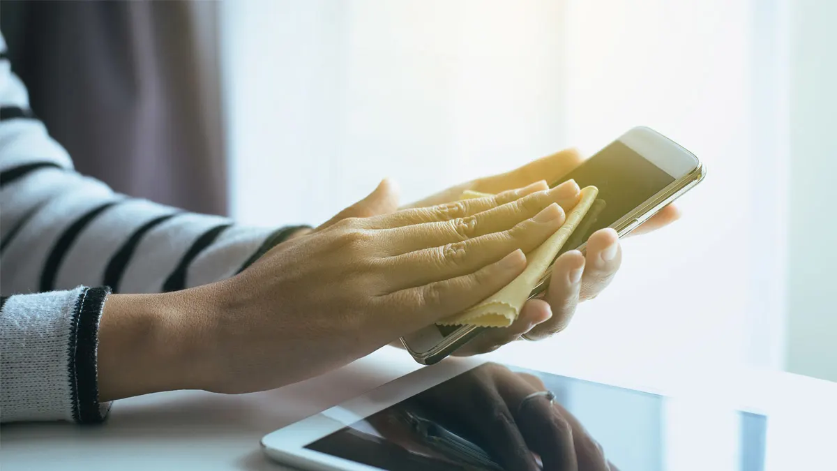 Eine Person wischt mit einem gelben Tuch über ein weißes Mobiltelefon. Auf dem Tisch darunter liegt ein Tablet.