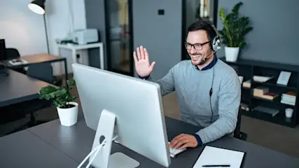 Ein Mann mit Headset winkt vor einem Computer an einem grauen Schreibtisch mit Pflanze und Notizblock. Im Hintergrund Büroausstattung.