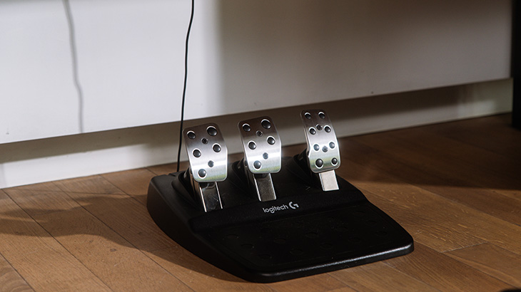 Drei silberne Pedale mit schwarzen Akzenten auf einer schwarzen Logitech-Basis, die auf einem Holzboden steht.