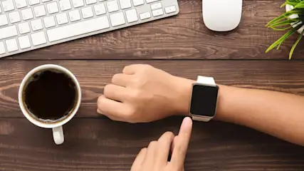 Auf einem Holztisch liegen eine Tastatur, eine Maus, eine Tasse Kaffee, eine Smartwatch am Handgelenk und eine Pflanze.