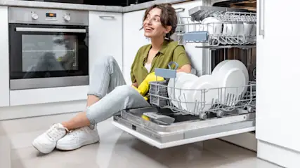 Eine Frau mit gelben Handschuhen sitzt vor einer offenen Spülmaschine in einer weißen Küche mit weißen Tellern und einem Ofen.
