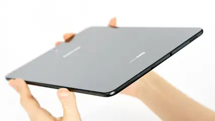Ein graues Tablet mit schwarzem Rand wird von zwei Fingern vor einem weißen Hintergrund gehalten.