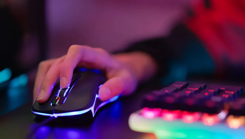 Eine Hand bedient eine beleuchtete schwarze Gaming-Maus auf einem dunklen Tisch, neben einer bunten Tastatur.