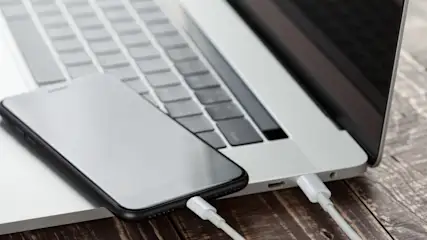 Ein Smartphone und ein Laptop liegen auf einem Holztisch. Beide Geräte sind mit einem weißen Kabel verbunden.