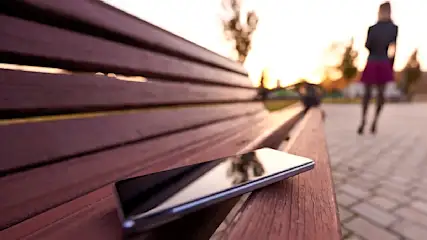 Ein Smartphone liegt auf einer braunen Holzbank. Im Hintergrund geht eine Person auf einem gepflasterten Weg.
