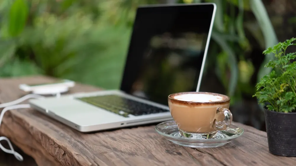 Ein Laptop mit Kabel, eine Tasse Kaffee auf Untertasse und eine kleine Pflanze stehen auf einem Holztisch.