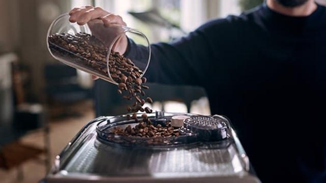 Eine Person gießt Kaffeebohnen aus einem Glasbehälter in eine Kaffeemühle. Bohnen fallen auf die silberne Oberfläche der Mühle.
