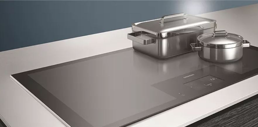 Edelstahltopf und Bräter auf einem schwarzen Induktionskochfeld mit weißer Arbeitsplatte vor einer blauen Wand.