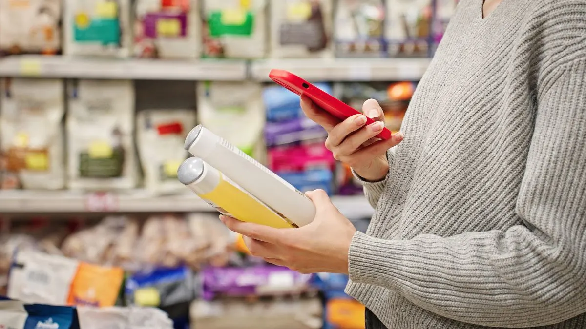 Eine Person steht in einem Laden. In der linken Hand hält sie Kosmetikprodukte, in der rechten ihr Smartphone, auf das sie schaut.