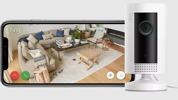 Ein Smartphone zeigt ein Wohnzimmer mit Sofa, Tisch und Stuhl, aufgenommen von einer Ring-Überwachungskamera, die daneben steht.