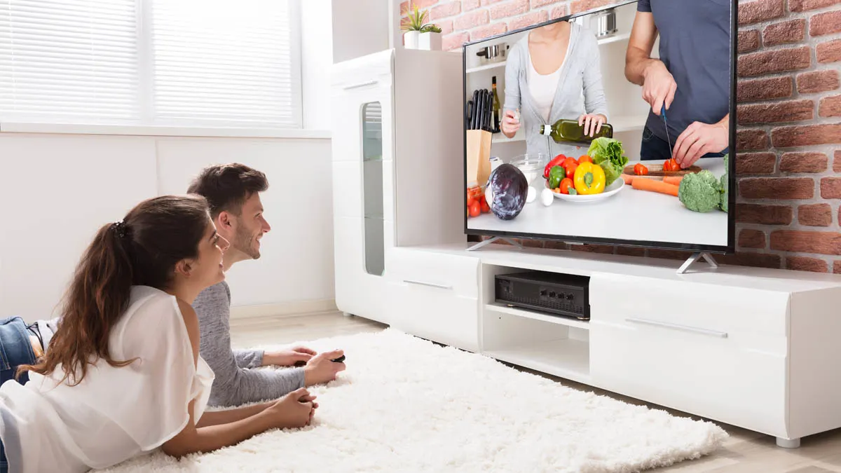 Eine Frau und ein Mann liegen auf einem weißen Teppich und schaut fern, mit einem Regal und einem Sofa im Hintergrund.