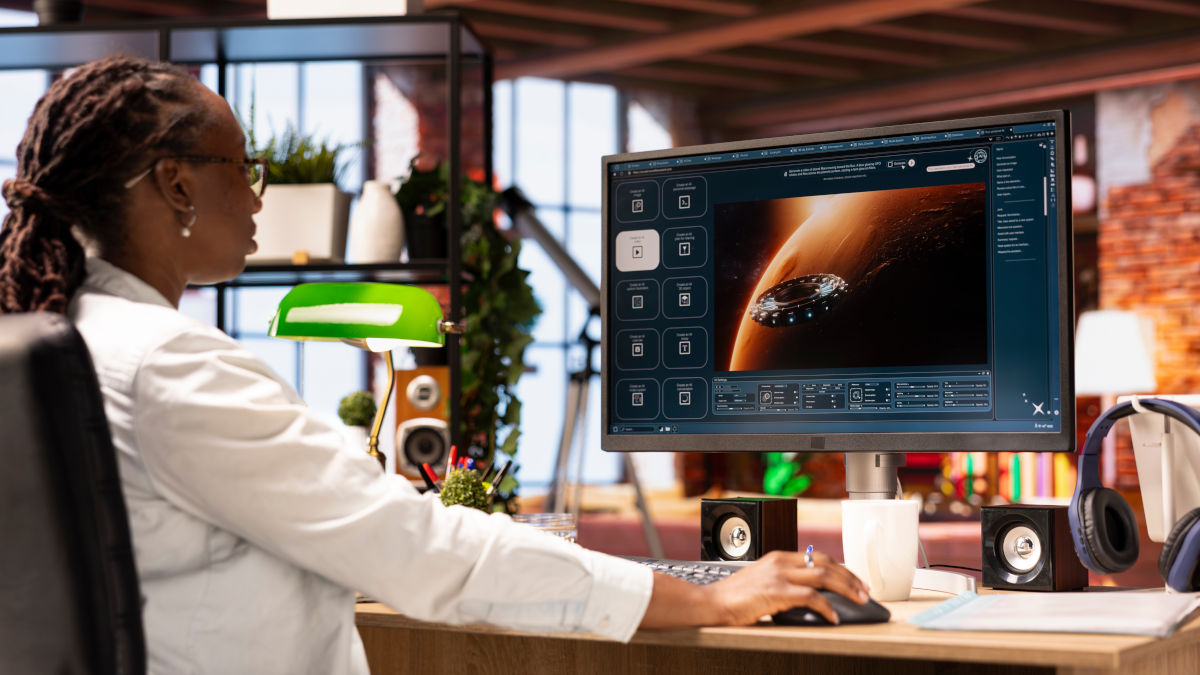 Eine Frau arbeitet am Computer mit Grafiksoftware. Lautsprecher und Kopfhörer sind auf dem Schreibtisch.