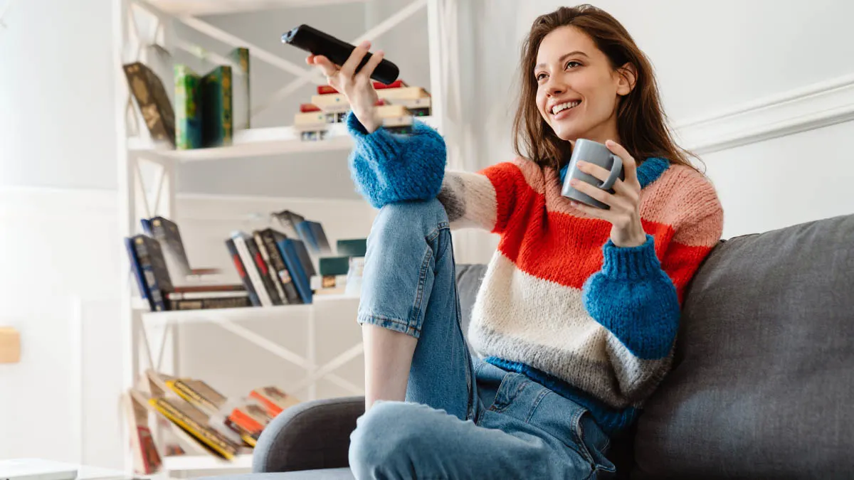 Eine Frau sitzt auf einer Couch und hält in der einen Hand eine Fernbedienung und in der anderen eine Tasse.