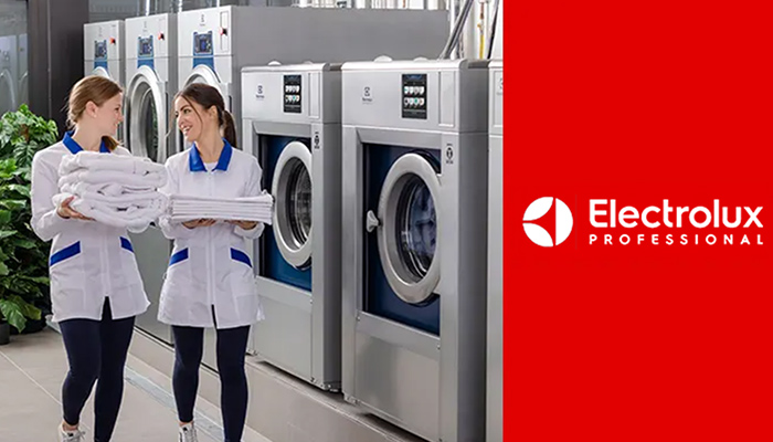 Zwei Frauen tragen je einen Stapel Wäsche, während sie an diversen Waschmaschinen vorbegehen.