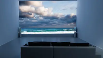Ein Raum mit Sofa blickt durch eine Öffnung auf das Meer mit Wolken am Himmel.