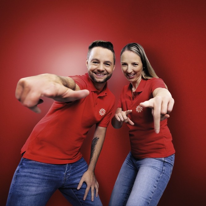 Zwei lächelnde Personen in roten Poloshirts zeigen vor rotem Hintergrund mit ausgestreckten Händen zur Kamera.