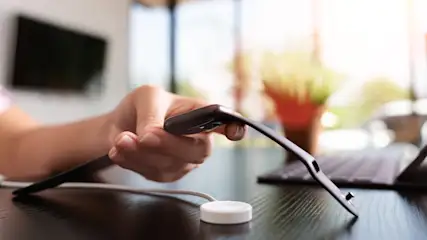 Eine Hand hält eine schwarze Smartwatch über ein weißes Ladegerät auf einem dunklen Tisch. Im Hintergrund sind ein Computer und eine Pflanze.